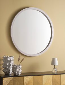 ein runder Spiegel in weißem Rahmen hängt an einer ockerfarbenen Wand, davor steht eine weiße Vase in gezackter Form und eine weiße kleine Lampe